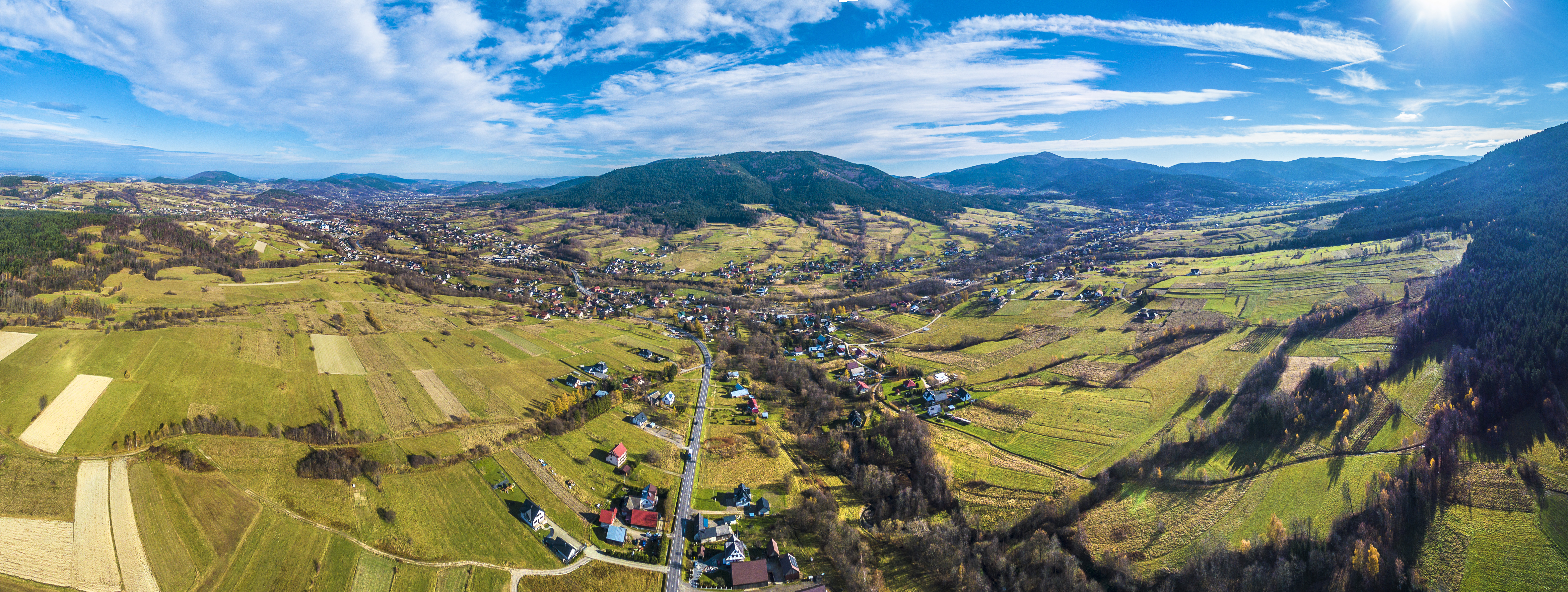 Panoramiczny widok na gminę Dobra, ukazujący malownicze tereny zielone, rozległe pola oraz zabudowania mieszkalne. Dominują pagórkowate krajobrazy, z drogą prowadzącą przez wiejskie osiedla, co podkreśla naturalne piękno okolicy i spokój regionu. Szerokie niebo z delikatnymi chmurami dopełnia całość tego harmonijnego krajobrazu.